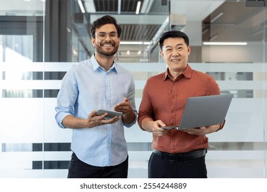 Two smiling businessmen stand in a modern office environment holding a tablet and laptop. Their casual attire complements the professional setting, indicating a collaborative atmosphere. - Powered by Shutterstock