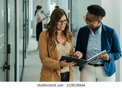 Two smiling businesswoman standing in a modern corridor and looking at new project on a papers while two unrecognizable coworkers talking in the background - Powered by Shutterstock