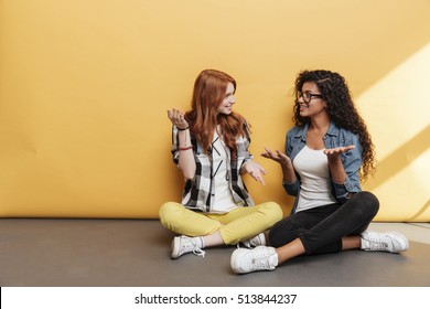 Two smiling confused young women sitting with legs crossed and talking over yellow background