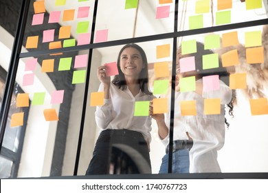 Two smiling diverse businesswomen planning project on papers, adding colorful post it notes with tasks or startup ideas on glass wall in office, coaches working with board together - Powered by Shutterstock