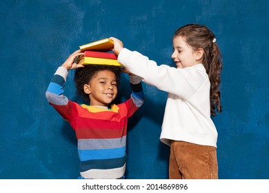 Two smiling diverse schoolchildren with textbooks, caucasian schoolgirl putting pile of books on head of mixed-race schoolboy on blue background. Back to school, children and education concept - Powered by Shutterstock