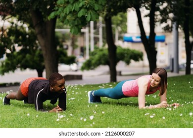 Two smiling diverse young woman in Athletic Workout Clothes are Doing a Plank Exercise working out together in park on grass.