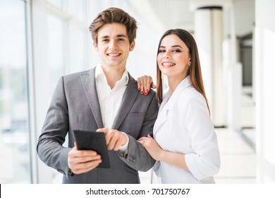 Two Smiling Elegant Business People Man In Formal Suit And Woman In Jacket And Dress Looking On Digital Tablet In Office Interior Near Window