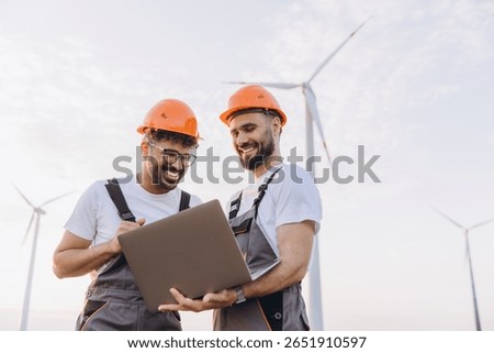 Two smiling engineers collaborating over a laptop, analyzing wind turbines for optimizing sustainable energy production and innovation