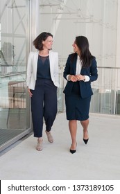 Two smiling female colleagues walking in office hall. Women chatting with glass walls in background. Break concept. Front view.