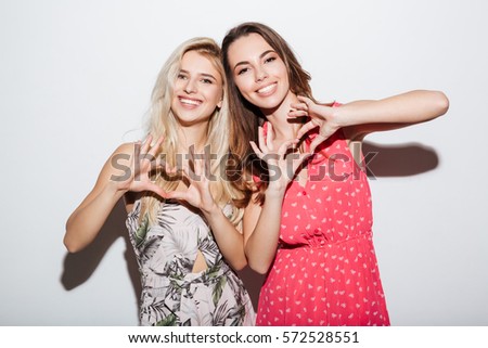 Two smiling girls in dresses showing heart gesture with hands isolated on the white background