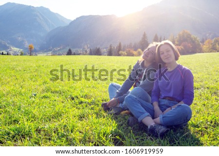 Two smiling girls are sitting on a green meadow and mountains in the background
