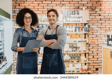 Two smiling helpful shop assistants standing close to goods shelves holding folder against brick wall. Multi-cultural sellers shop owners cafeteria staff waiters posing for camera - Powered by Shutterstock