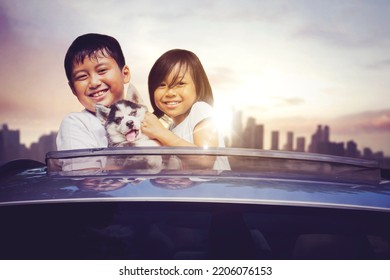 Two Smiling Kids Embracing Their Dog On The Sunroof Of Car While Enjoying Road Trip With Dusk Sunlight Background