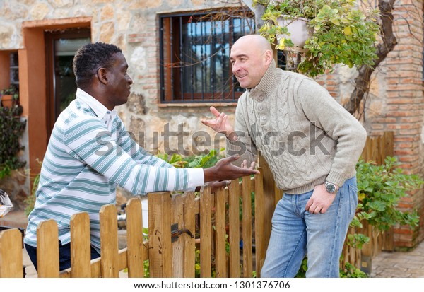 Two smiling male neighbors talking\
through wooden fence, discussing latest\
news\
\
