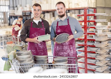 Two smiling male potters in stained maroon aprons holding handcrafted ceramic plates, conveying sense of accomplishment in artisanal pottery workshop - Powered by Shutterstock