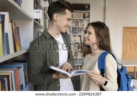 Two smiling pretty girl and guy with textbook standing in library, prepare for exams together, discuss exercise, joint task or essay, talking during meeting indoors. Teamwork, education, friendship