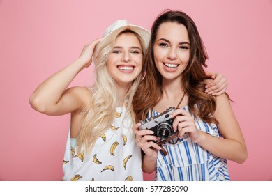 Two smiling pretty young women with old vintage photo camera over pink background