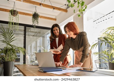 Two smiling professional female partners or coworkers, happy business women entrepreneurs working together in office looking at laptop using computer writing notes standing at work desk.