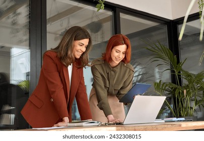 Two smiling professional female partners or colleagues, happy business women entrepreneurs working together in office looking at laptop using tablet computer technology standing at work desk. - Powered by Shutterstock