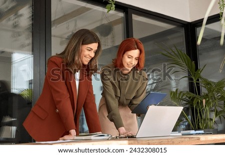 Two smiling professional female partners or colleagues, happy business women entrepreneurs working together in office looking at laptop using tablet computer technology standing at work desk.