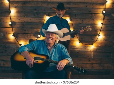 Two smiling senior country and western musicians sitting on chair in front of wooden wall with light bulbs.