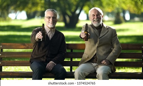 Two smiling senior friends showing beer bottle into camera, relaxing in park