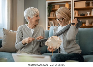 Two smiling senior women enjoying retirement, sitting on a cozy sofa in their living room, knitting a gray scarf and sewing, sharing creativity and friendship