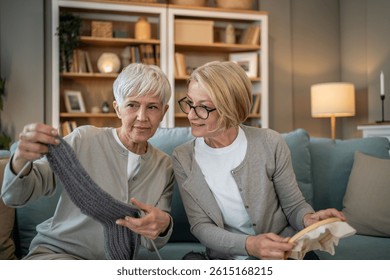Two smiling senior women enjoying their retirement, sitting on a comfortable sofa in their living room, knitting and embroidering together, sharing a moment of relaxation and creativity