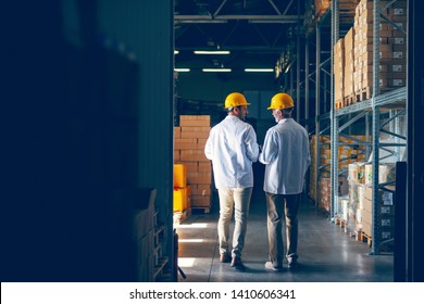 Two Smiling Warehouse Workers In White Uniforms And Yellow Helmets On Heads Standing And Talking About Job. Backs Turned.