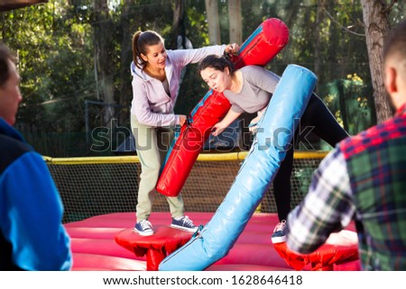 Two smiling women having funny battle by big stuffed beams on inflatable arena at amusement park