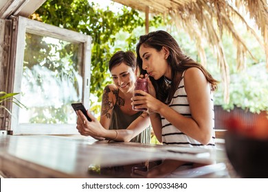 Two Smiling Women Sitting At A Restaurant Looking At Mobile Phone. Woman Showing Mobile Phone While Another Woman Enjoys A Smoothie.