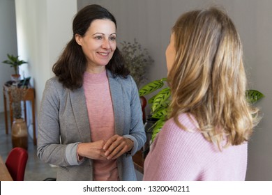 Two Smiling Women Standing And Talking. Woman Wearing Casual Clothes And Talking With Her Colleague Or Client Who Is Standing Back To Camera With Office Interior In Background. Conversation Concept.