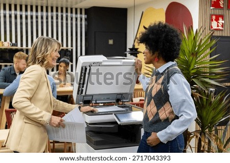 Two smiling women using a printer in a modern office, showing teamwork and collaboration among diverse colleagues