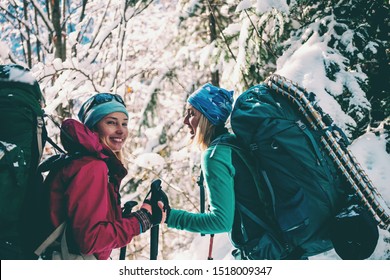 Two smiling women in a winter hike. Girlfriends with trekking poles are on a snow covered mountain path. Girls with backpacks and snow shoes travel together. Friends walk through the fir forest.