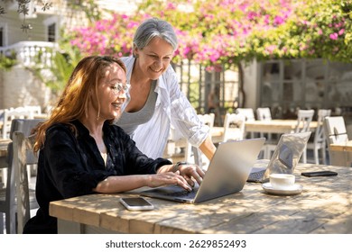Two smiling women work together on laptops at an outdoor café. Surrounded by flowers and sunlight, they represent teamwork, freelance collaboration, and a modern remote work lifestyle.