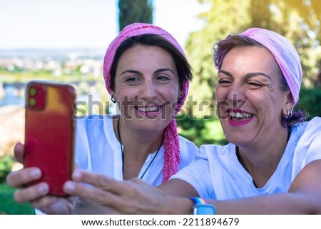 Two smiling young adult women with a pink headscarf taking a selfie. 2 warrior sisters laughing and happy after overcoming breast cancer. Breast Cancer Awareness Day concept. Blur background