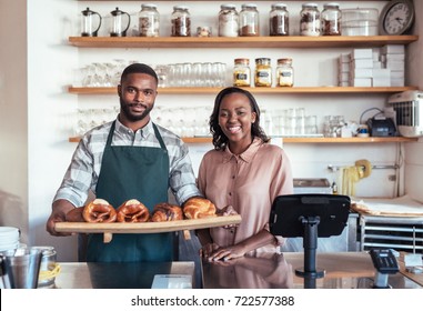 Two smiling young African entrepreneurs holding a tray of baked goods while standing behind the counter of their bistro - Powered by Shutterstock