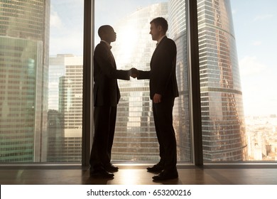 Two smiling young black and white businessmen handshaking standing near big window with city buildings outside, confident caucasian and afro american partners form good relations, reach agreement
