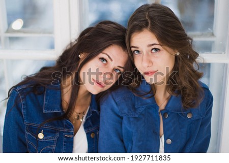 Two smiling young brunette twins sisters girls. Portrait shot of two gorgeous women in blue jeans jackets with perfect look, posing. Family concept. Cute and intelligent looking girls. High fashion 