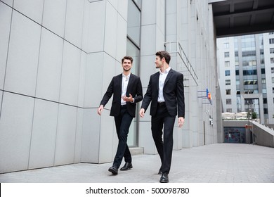 Two Smiling Young Businessmen Walking And Talking In The City