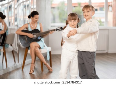 Two smiling young children, girl and boy engaging in ballroom dance lesson while female instructor providing live guitar music in bright choreography studio .. - Powered by Shutterstock