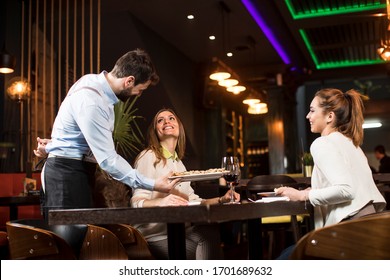 Two Smiling Young Female Friends At A Restaurant With Waiter Serving Dinner