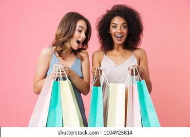 Two Smiling Young Girls Dressed In Summer Clothes Looking Inside Shopping Bags Isolated Over Pink Background