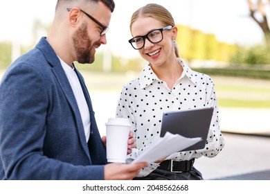 Two Smiling Young Man And Woman Business Colleagues Dressed In Formal Clothes Discussing Work While Standing Outsisde With Digital Tablet And Documents In Hands, Cheerful Coworkers Talking Outdoors