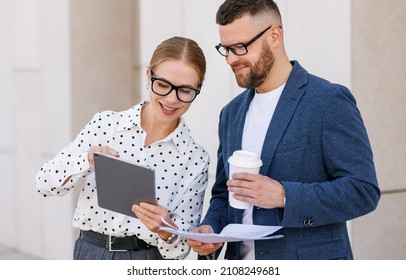 Two Smiling Young Man And Woman Business Colleagues Dressed In Formal Clothes Discussing Work While Standing Outsisde With Digital Tablet And Documents In Hands, Cheerful Coworkers Talking Outdoors