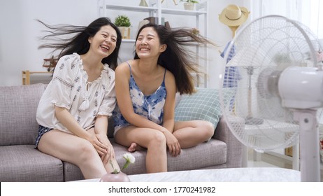 Two Smiling Young Woman Friends Blowing Hair In Wind From Electric Fan. Beautiful Girls In Shorts In Hot Summer Day Stay At Home Sitting On Sofa Laughing Gossip Talking Chatting With Air Cooler.