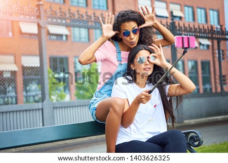 Two smiling young women making faces while sitting on a city bench taking self portraits together with a smartphone and selfie stick