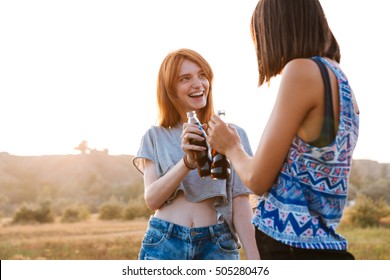 Two Smiling Young Women Standing And Drinking Soda Outdoors