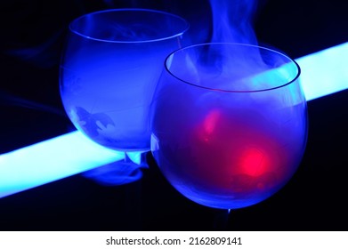 Two Smoky Cocktails With A Neon Tube, On A Dark Background. One Of The Glasses Slowly Igniting Alcohol Inside. Dangerous Liquor Mixture - Close Up Photo. Background Picture.