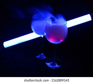 Two Smoky Cocktails With A Neon Tube, On A Dark Background. One Of The Glasses Slowly Igniting Alcohol Inside. Dangerous Liquor Mixture - Close Up Photo. Background Picture.