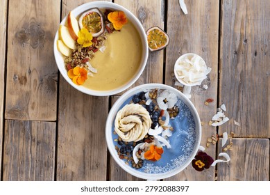 Two smoothies sit on a rustic wooden table. One has apple slices and passion fruit toppings. The other features coconut and berry decorations. Edible flowers and seeds adorn both bowls