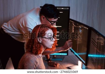 Two software developers collaborate at dual monitors, reviewing code late in a dim office. Coding business concept