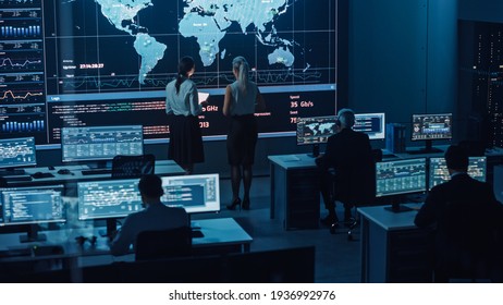 Two Software Engineers Talking Next to a Big Screen in a Modern Monitoring Office with Live Analysis Feed with Charts. Monitoring Room Big Data Scientists and Managers Sit in Front of Computers.