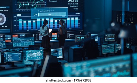 Two Software Engineers Talking Next to a Big Screen in a Modern Monitoring Office with Live Analysis Feed with Charts. Monitoring Room Big Data Scientists and Managers Sit in Front of Computers. - Shutterstock ID 1936992982